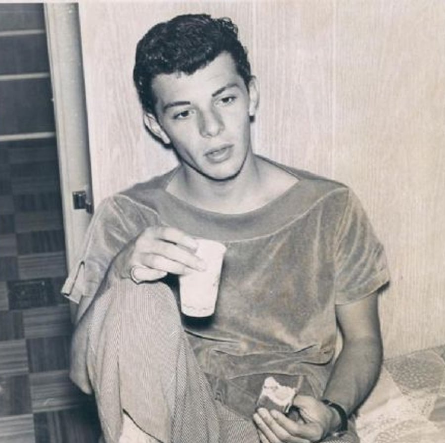 Young man with short, dark hair sits casually against a wall, holding a cup in one hand and a piece of food in the other. He wears a loose t-shirt and checkered pants, looking slightly to the side with a relaxed expression.