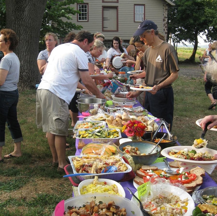 People are serving themselves food from a long table filled with various dishes at an outdoor picnic or potluck gathering. There are trees and a house in the background, and the atmosphere seems casual and friendly.