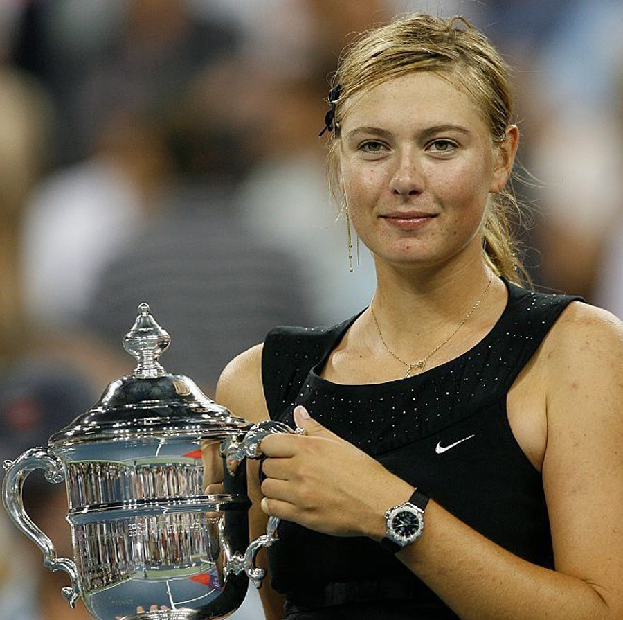 A female tennis player in a black dress holds a large silver trophy and smiles slightly. She is standing on a tennis court, with a blurred crowd in the background.