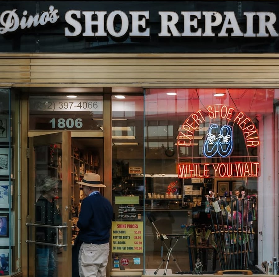 Storefront of "Dino's Shoe Repair" with a neon "Expert Shoe Repair While You Wait" sign in the window. Two people stand at the entrance, and various shoe care items are visible inside.