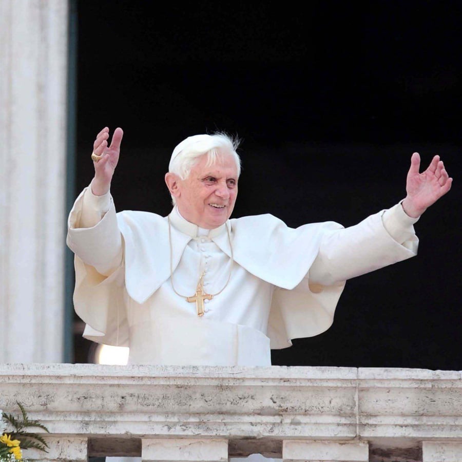 An elderly man dressed in white religious robes and a cross necklace stands on a balcony with his arms raised, smiling at a crowd below.