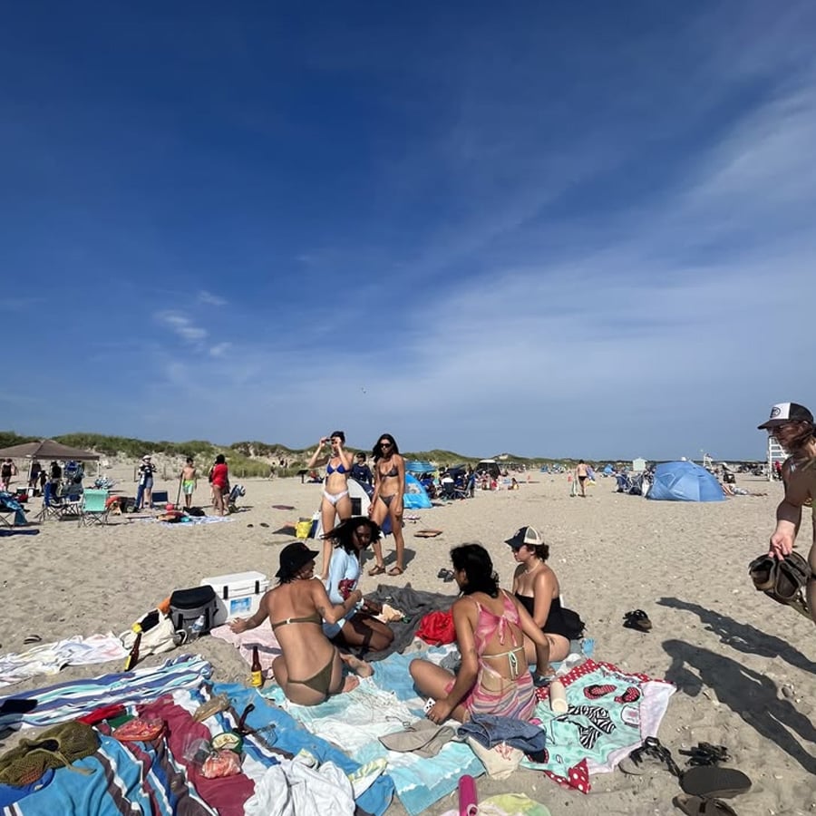 A group of people in swimsuits sit and stand on colorful towels and blankets at a sandy beach, with others and tents in the background under a blue sky.