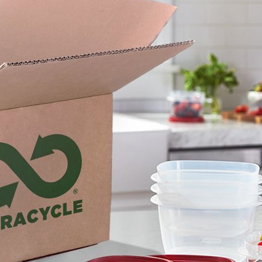 A cardboard box with a recycling symbol and the word “RECYCLE” printed on it sits on a kitchen counter near stacked plastic containers and fresh produce in the background.