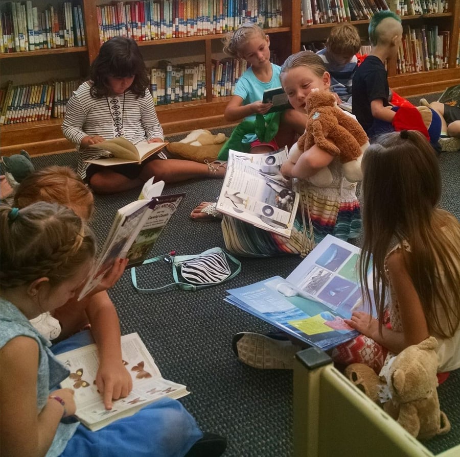 A group of young children sit on the floor in a library, reading books together. Some are holding stuffed animals, and bookshelves filled with books are visible in the background.