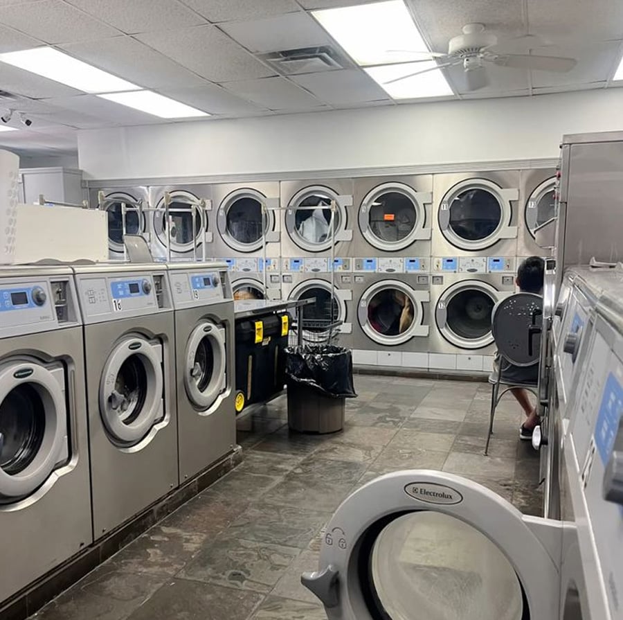 A laundromat with rows of front-loading washers and dryers; one person sits on a chair near laundry carts and baskets, waiting. The room is well-lit with white walls and a tiled floor.