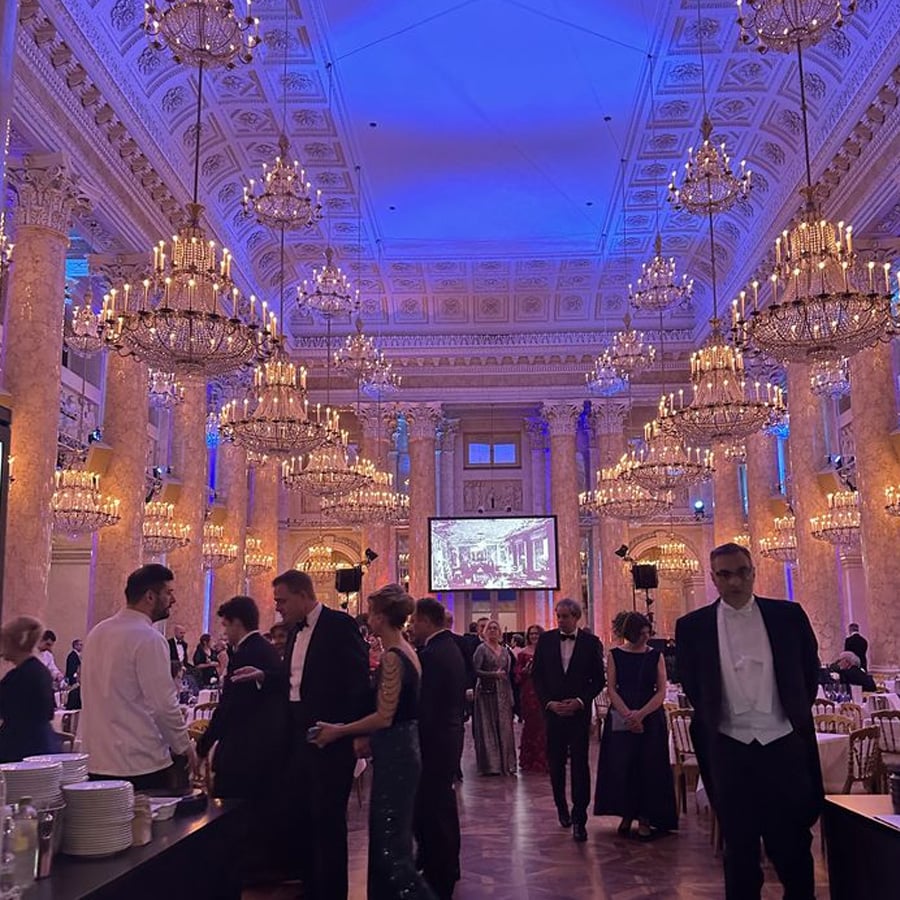 Guests in formal attire mingle in an opulent ballroom with marble columns, ornate ceilings, and grand chandeliers. Tables are set for dinner, and a large screen displays an image at the far end of the room.