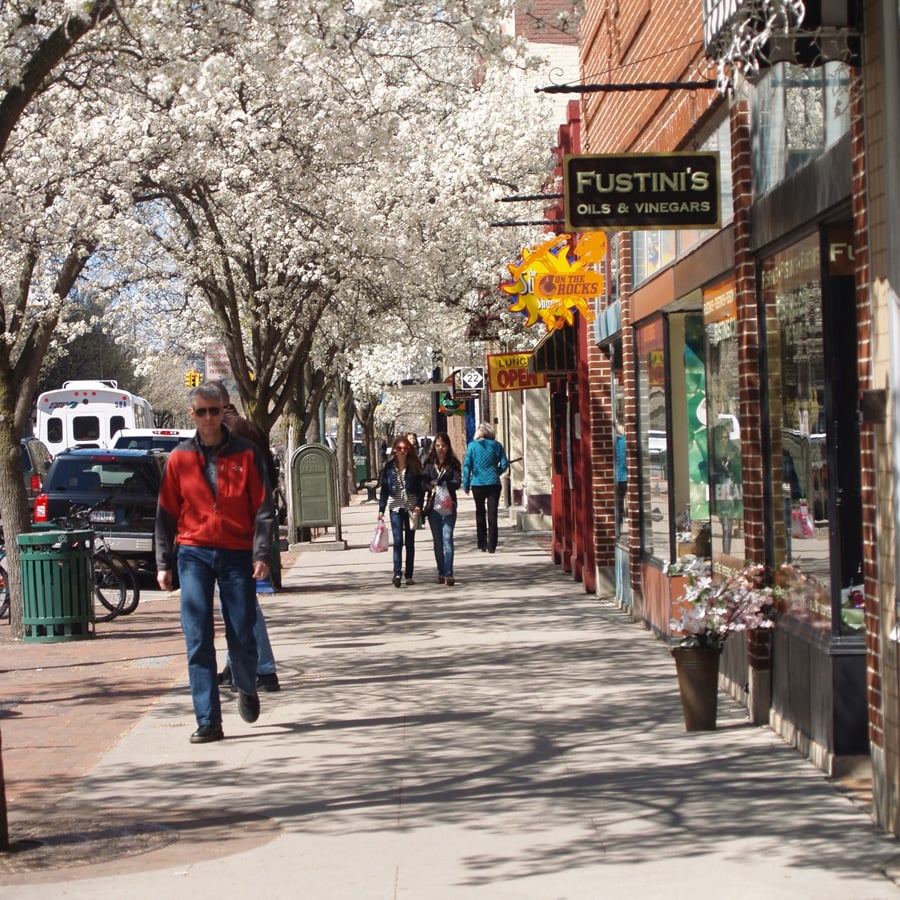 People walk along a sunny sidewalk lined with blooming trees and brick storefronts. Signs for shops like "Fustini's Oils & Vinegars" and "Sweet Shop" are visible. An ambulance is parked on the street.