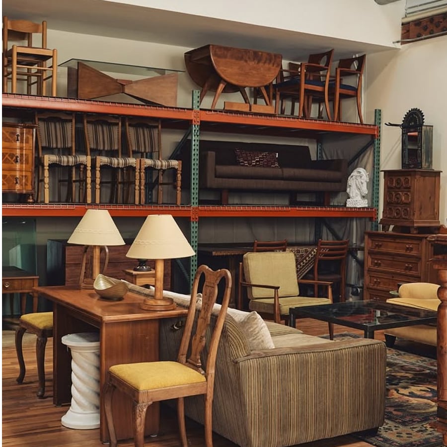 A room filled with vintage furniture, including wooden chairs, tables, sofas, lamps, and shelves. Several items are stacked or displayed on metal racks against the wall, creating an eclectic and cozy atmosphere.