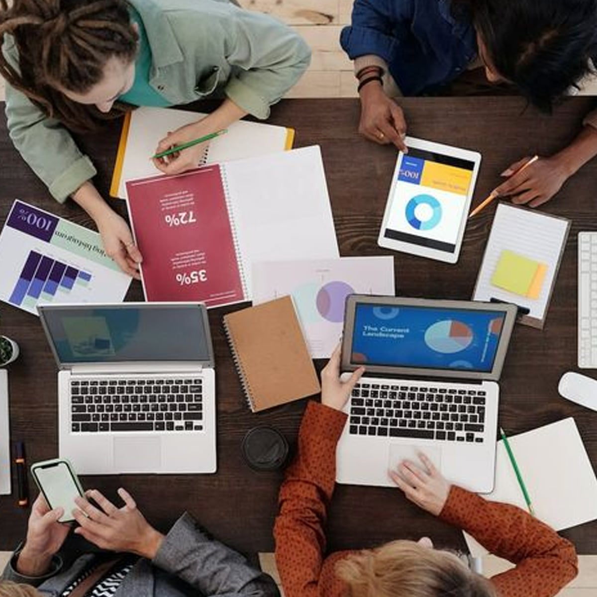 Four people work together at a table with laptops, tablets, notebooks, graphs, and charts, collaborating on a project with colorful documents and digital devices spread out in front of them.