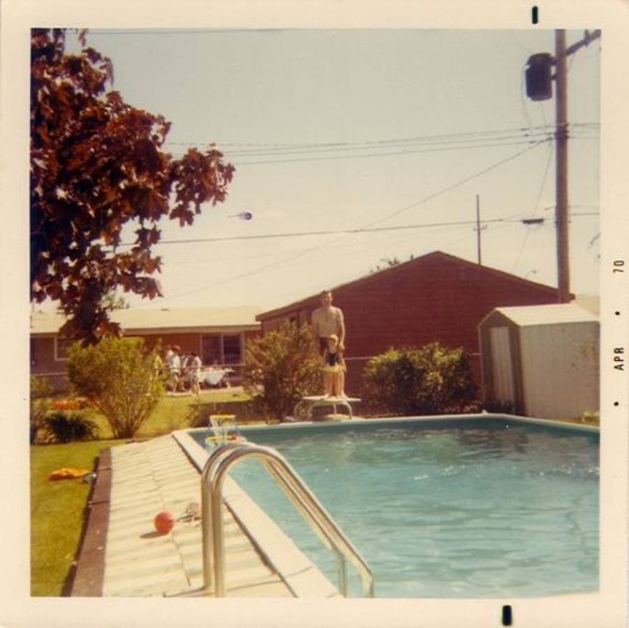A vintage photo of a backyard pool with a man standing on a diving board. There’s a tree, bushes, and a fence nearby; a house and several people are visible in the background. The photo is dated April 1970.