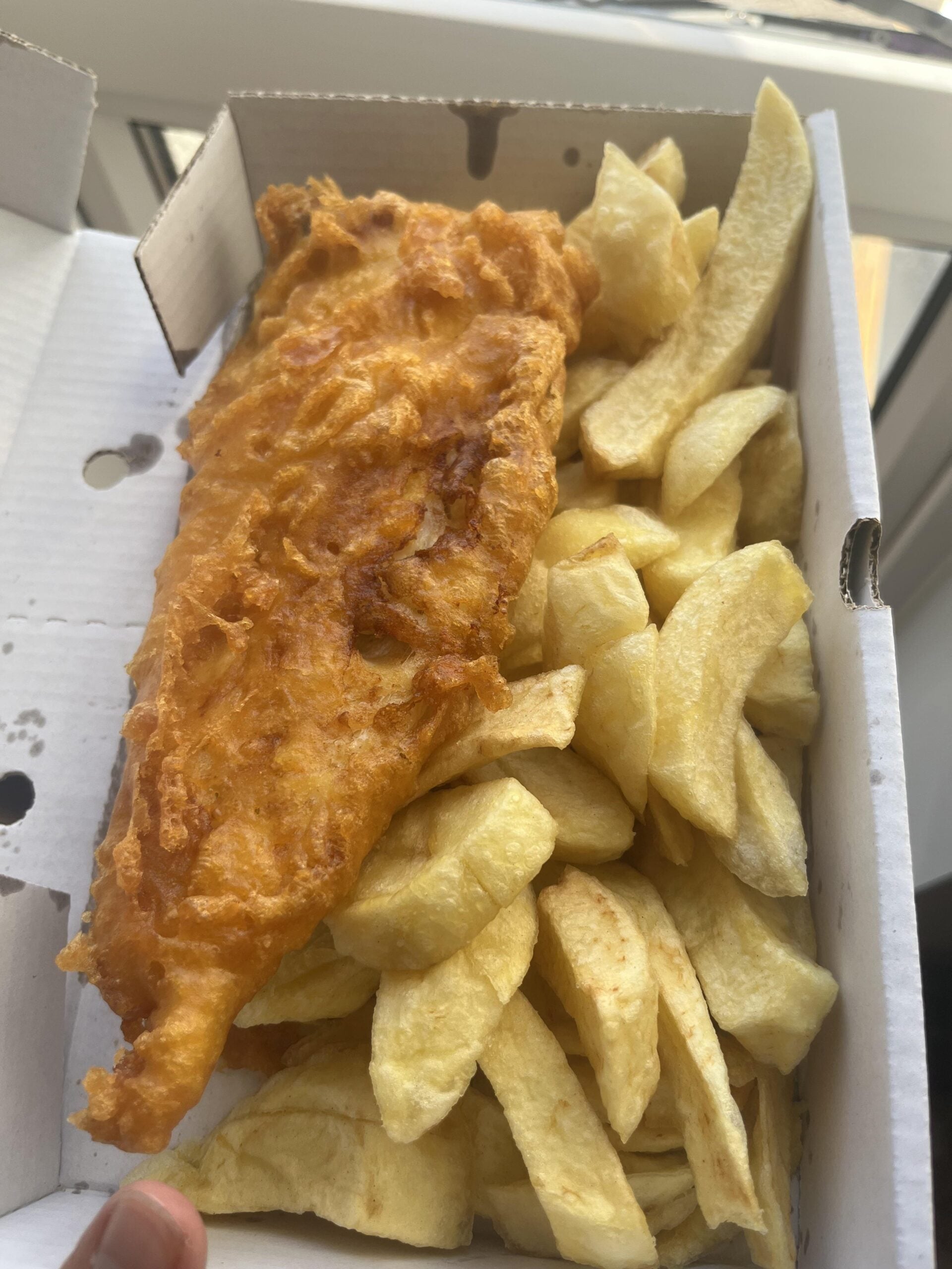 A portion of battered and fried fish served with thick-cut chips in a white cardboard takeaway box, placed near a window with natural light.