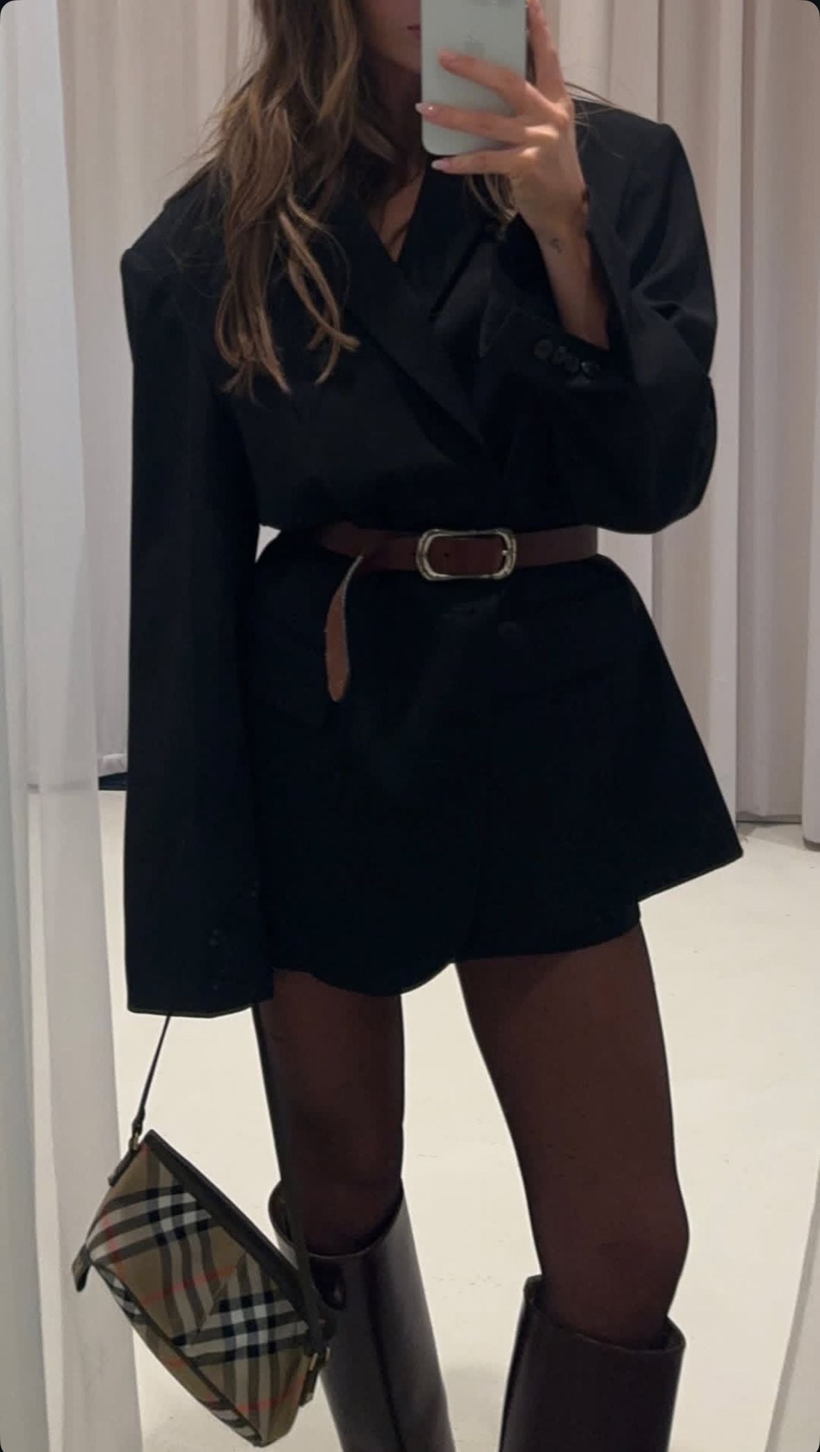 A woman takes a mirror selfie wearing an oversized black blazer as a dress, belted at the waist, with black shorts, dark tights, knee-high boots, and holding a plaid handbag. Her face is not visible.