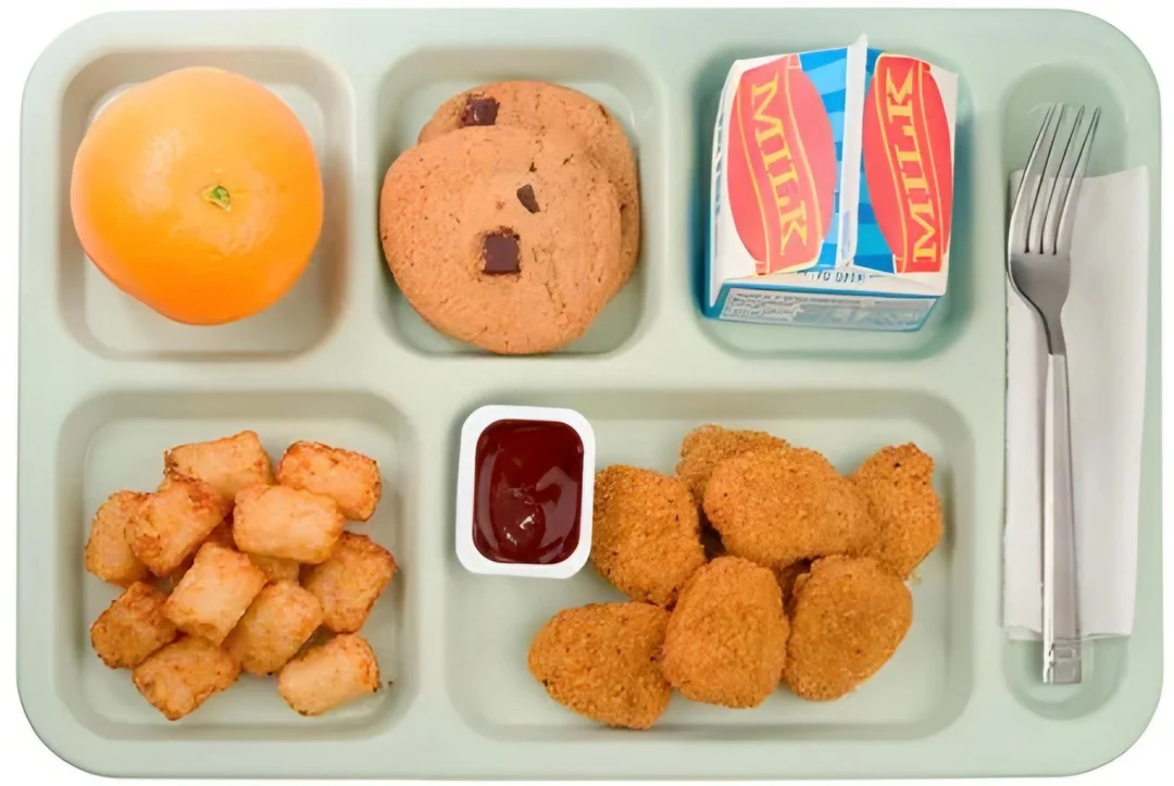 A school lunch tray with an orange, two chocolate chip cookies, a carton of milk, tater tots, a serving of ketchup, chicken nuggets, and utensils wrapped in a napkin.