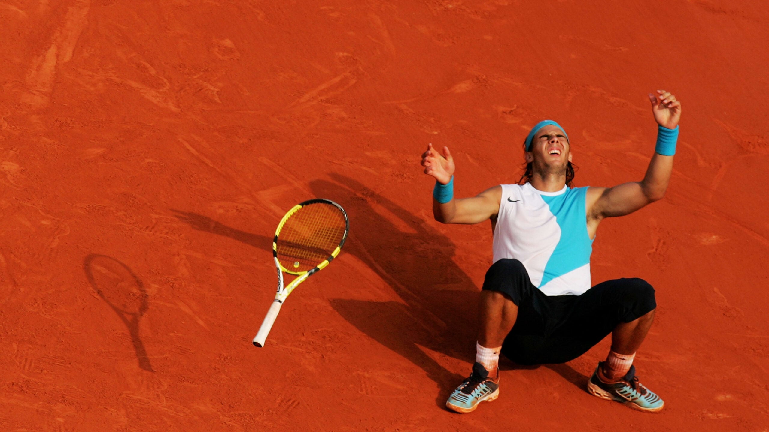 A tennis player wearing a blue and white outfit sits on a clay court with arms raised and eyes looking up, celebrating. His racket lies on the ground nearby, casting a shadow on the orange surface.