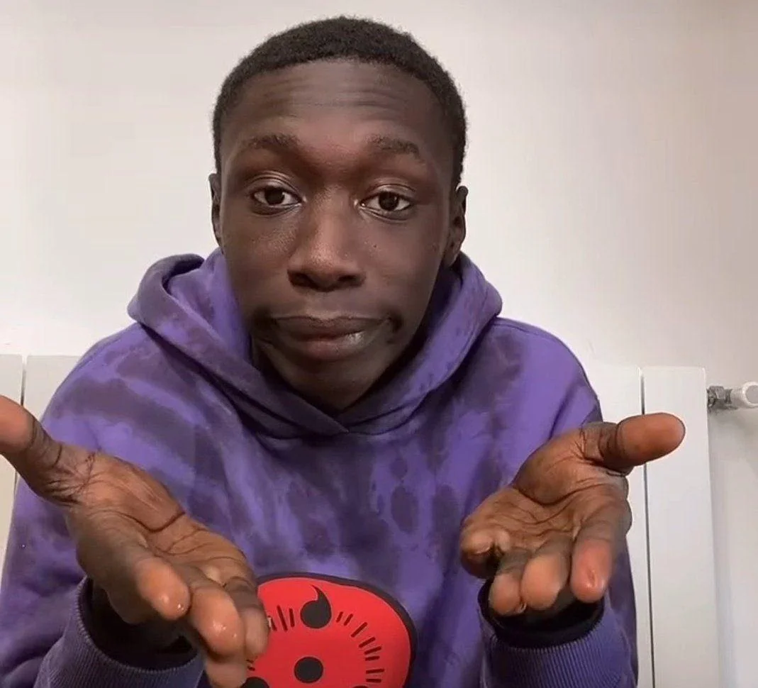A person wearing a purple hoodie with a red design on the front holds their hands out with an expression of confusion or questioning, as if asking “why” or “what’s going on.”