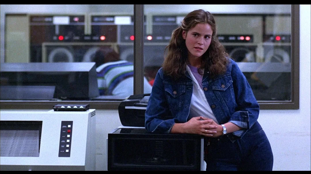 A young woman with long brown hair, wearing a denim jacket and jeans, leans against a desk in a room with vintage computers and large machines in the background. She looks thoughtfully toward the camera.