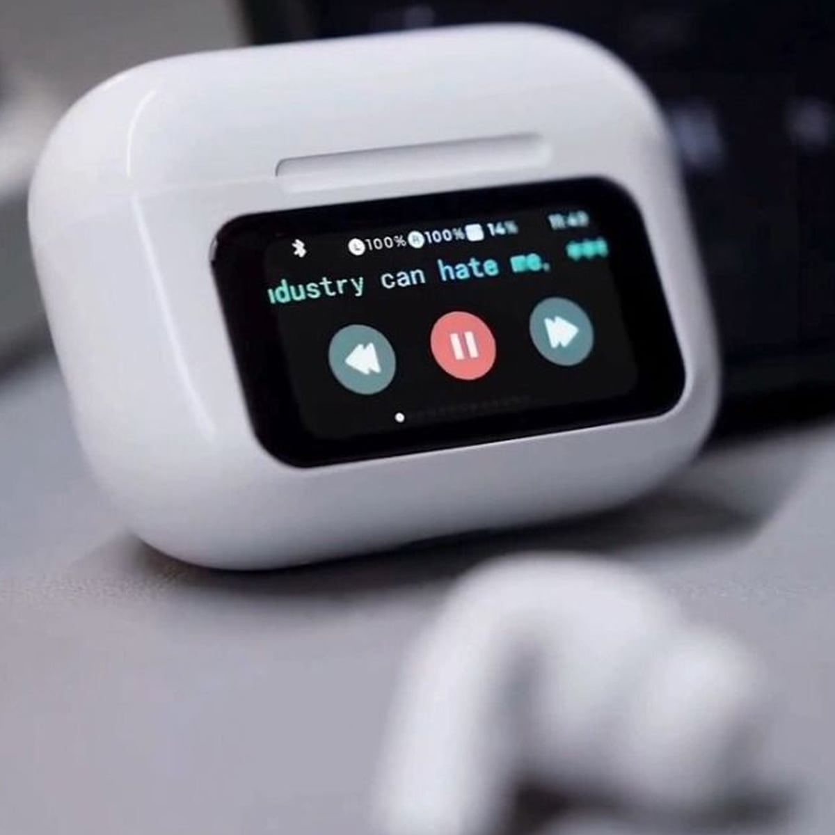 A close-up of a white wireless earbud case with a small screen displaying music playback controls and partial lyrics, "dustry can hate me," with play, pause, and forward buttons visible. An earbud rests in the foreground.