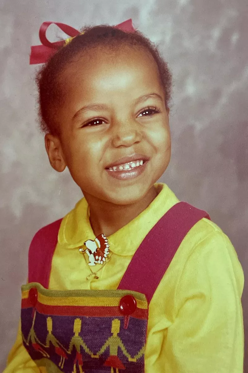 A young child with a big smile wears a yellow shirt under red overalls with a colorful pattern and a necklace. She has a red bow in her hair and poses in front of a gray studio backdrop.