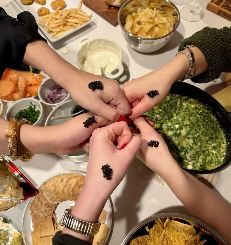 Five hands with black caviar on them form a circle above a table filled with various foods, including chips, crackers, dips, and a green spinach dip. The scene suggests a festive or social gathering.