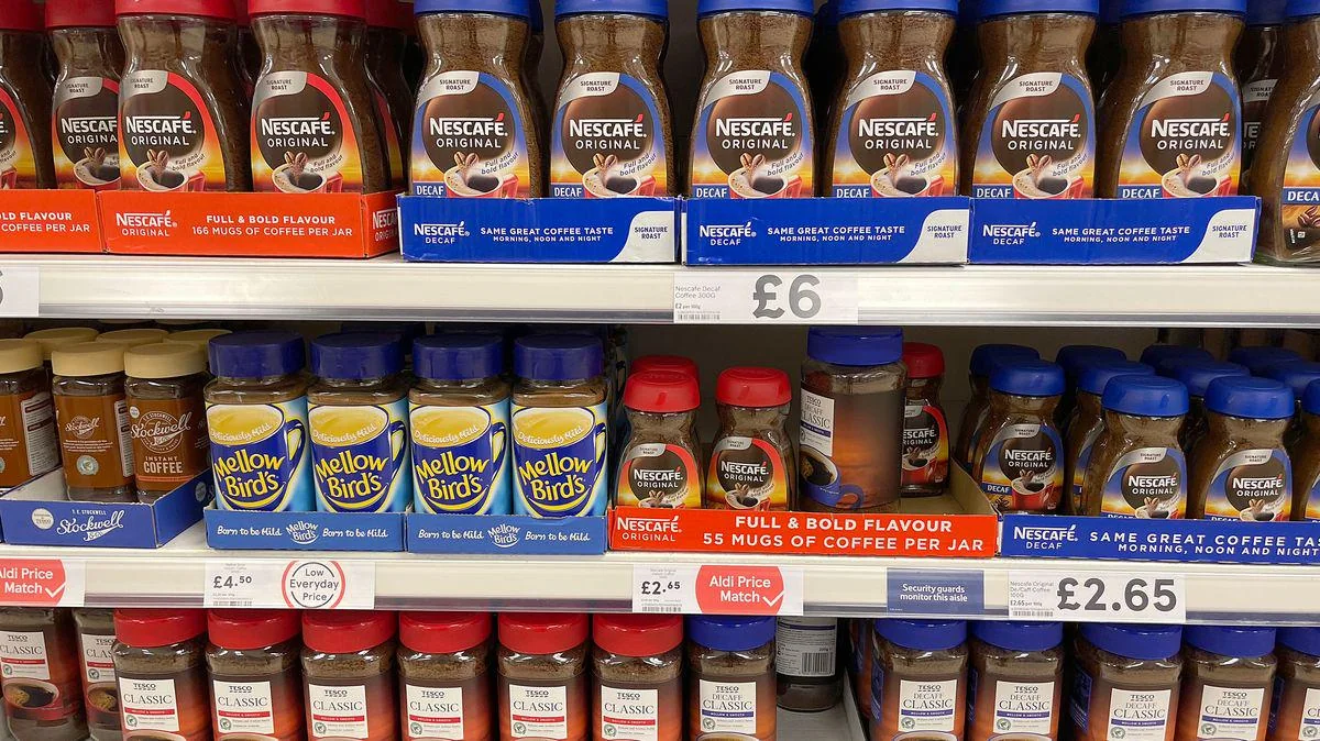 Shelves in a supermarket display Nescafé and Yellow Birds instant coffee jars in various sizes, with price tags below each row. The jars have red, blue, or yellow lids and colorful labels.