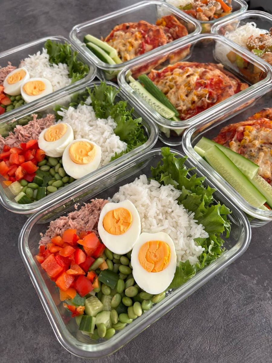 Glass meal prep containers filled with rice, boiled egg halves, lettuce, tuna, edamame, red bell pepper, and cucumber slices. In the background, containers with baked dishes topped with melted cheese are visible.