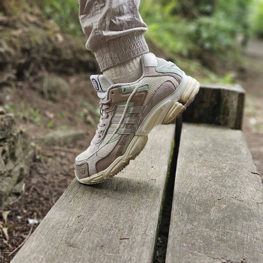 A person wearing beige sneakers and light pants steps onto a wooden bench outdoors, surrounded by greenery and a dirt path.