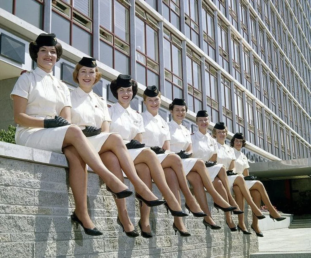 Eight women in matching white uniforms, black hats, gloves, and heels sit in a row on a low wall outside a modern building, smiling and posing with their legs crossed and facing the camera.