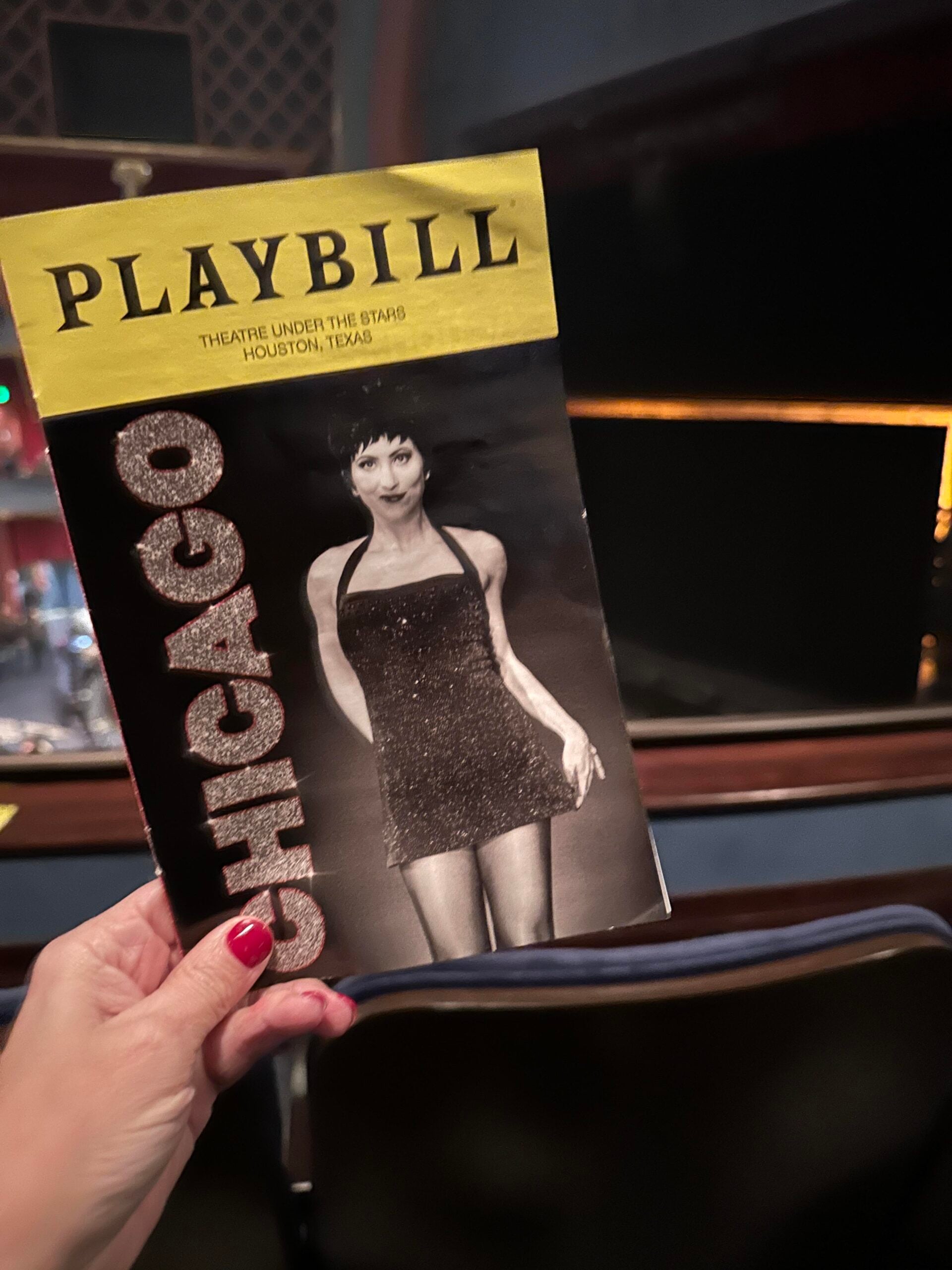A hand holds a Playbill for the musical "Chicago" at Theatre Under The Stars in Houston, Texas, featuring a performer in a sparkly black dress on the cover. Theater seats and stage are visible in the background.