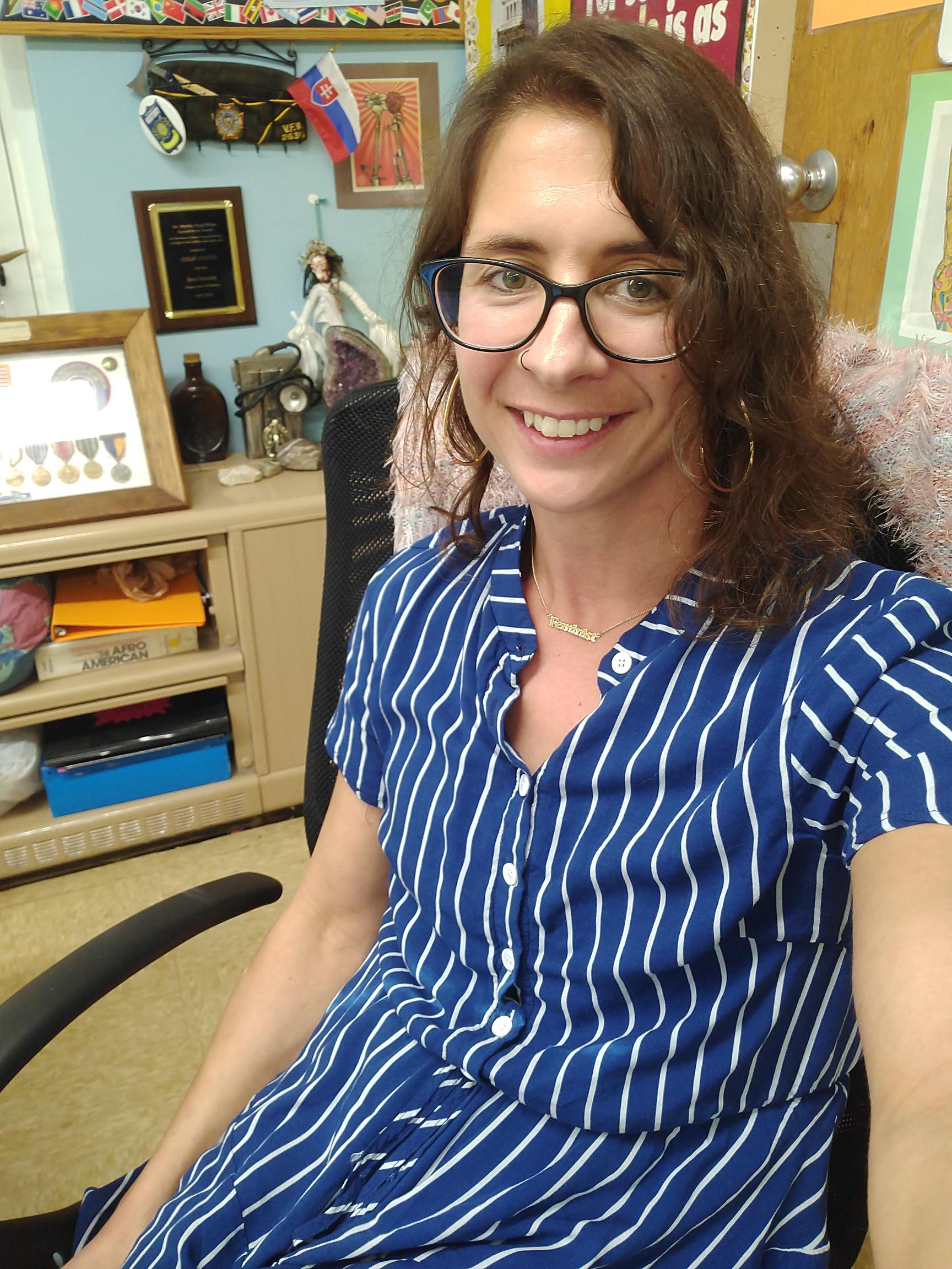 A woman with wavy brown hair, glasses, and a blue-and-white striped dress smiles while sitting in an office chair. Behind her are colorful decorations, awards, and educational materials on the wall and shelves.