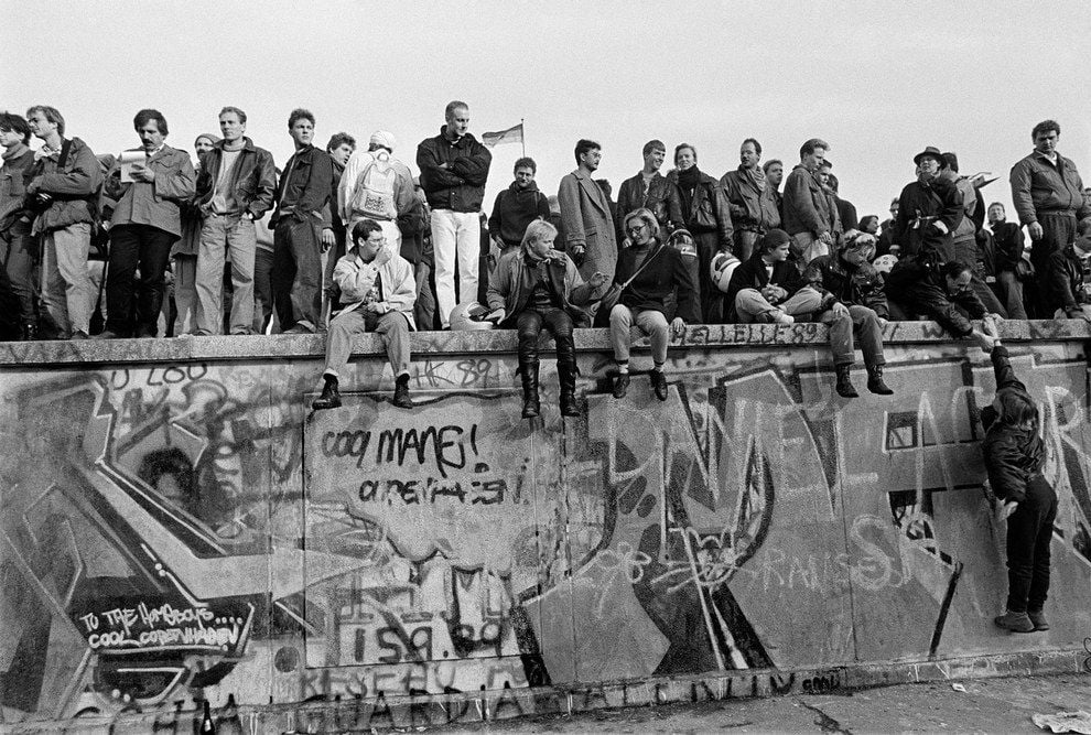 A large group of people stands and sits atop a graffiti-covered concrete wall, gathered closely together, watching and interacting, in what appears to be a historic moment. The scene is in black and white.