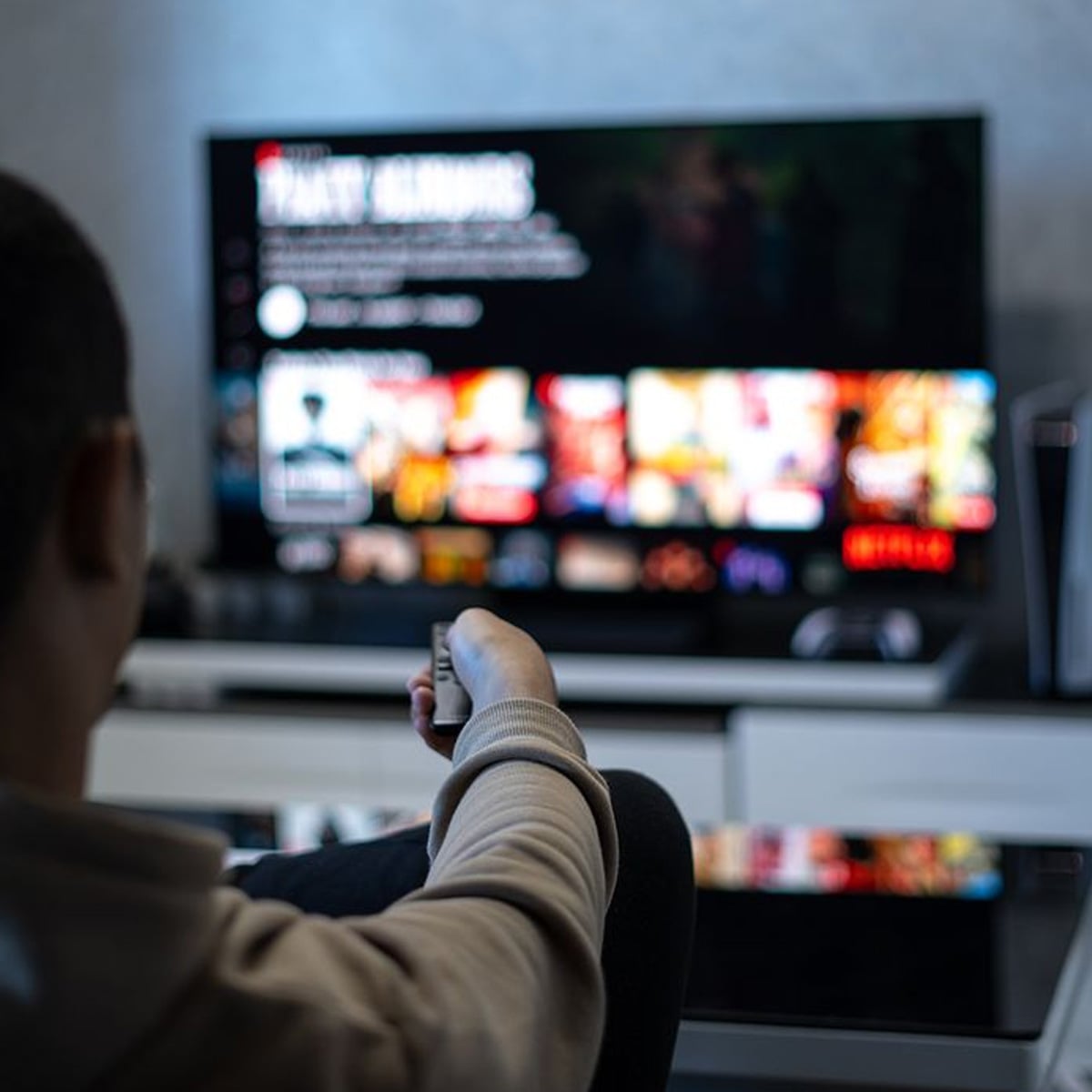 A person sitting on a couch, holding a remote control and facing a TV screen displaying a streaming service interface with various movie and show thumbnails.