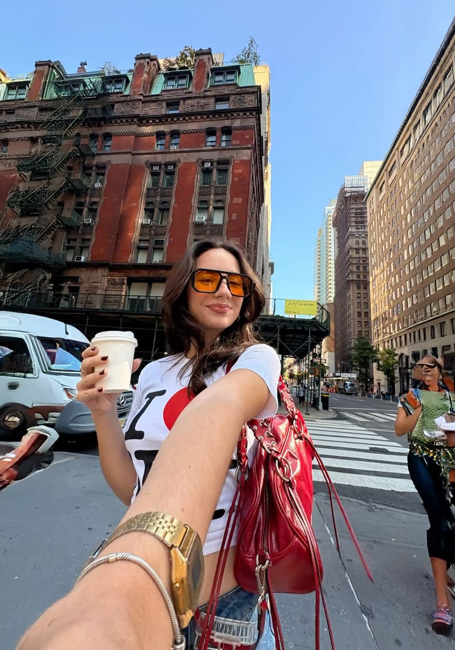 A woman wearing sunglasses and an "I ❤️ NY" shirt takes a selfie on a city street. She holds a coffee cup and a red bag, with tall buildings and people in the background on a sunny day.