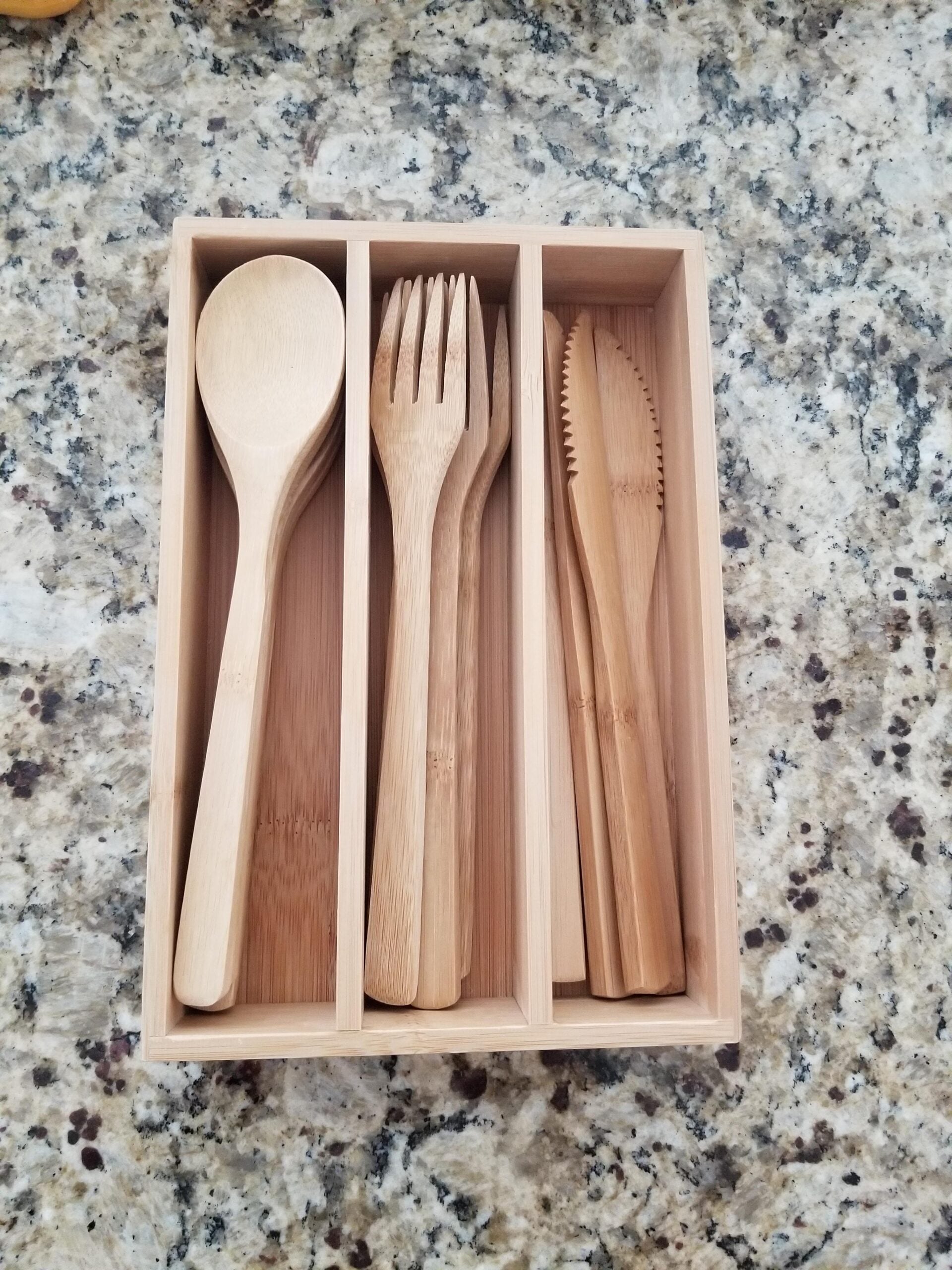 A wooden utensil tray holds bamboo spoons, forks, and knives, neatly organized in separate sections, placed on a speckled kitchen countertop.