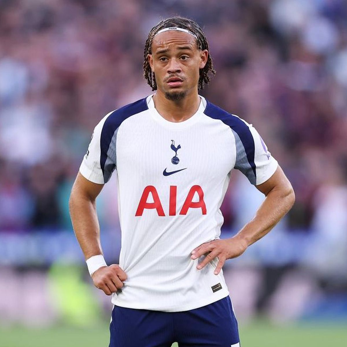 A football player in a white and navy Tottenham Hotspur kit stands on the field with his hands on his hips during a match, looking focused. The crowd and stadium are blurred in the background.