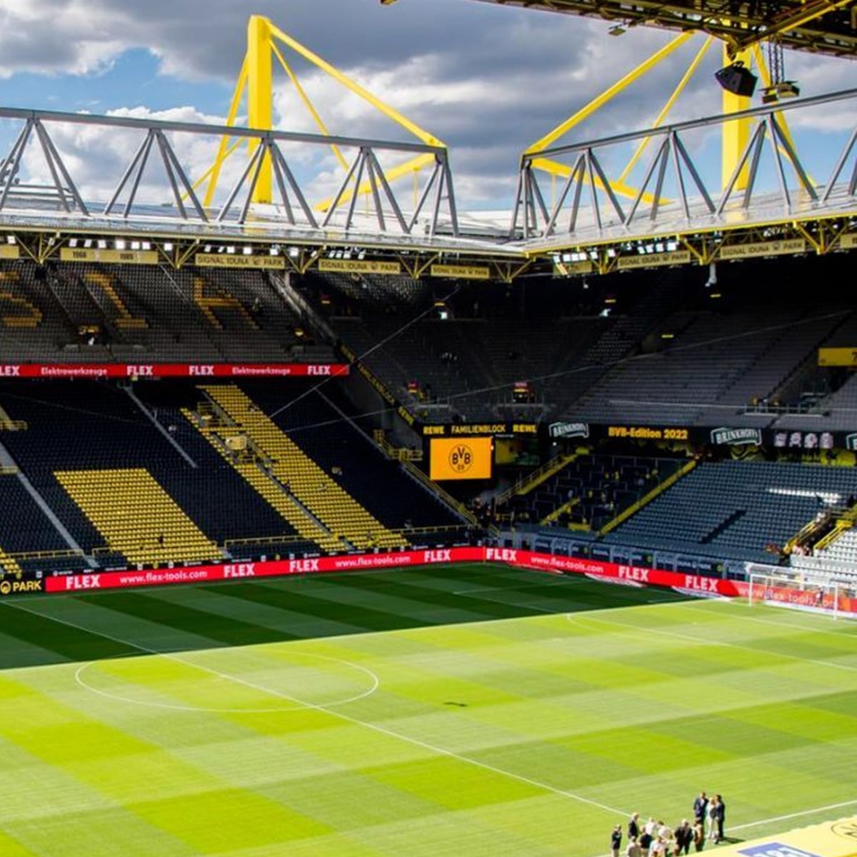 A large football stadium with yellow and black seating, modern architecture, and a green pitch under a partly cloudy sky. A few people stand near the corner of the field.