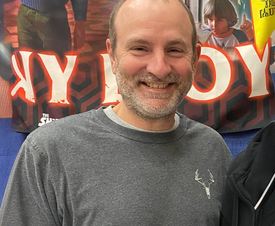 A smiling man with short hair and a beard wearing a gray shirt with a deer logo stands indoors; behind him is a colorful banner with large red letters and a partially visible child.