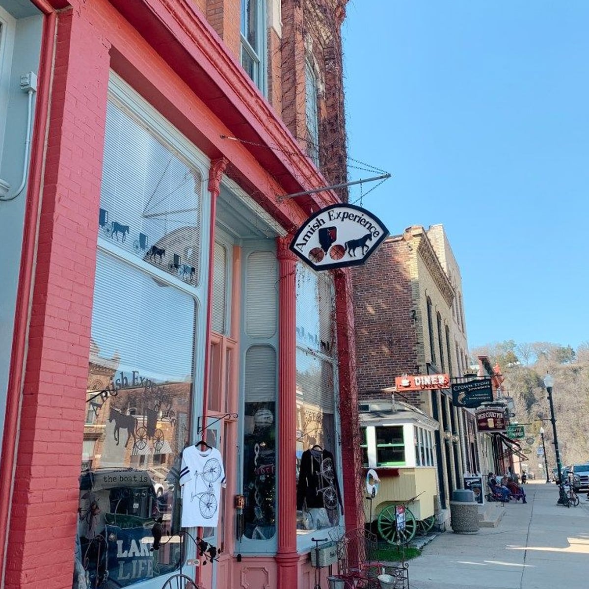 A street scene shows the front of a shop called "Amish Experience" with a hanging sign, brick buildings, and a few people and benches along the sidewalk on a sunny day.