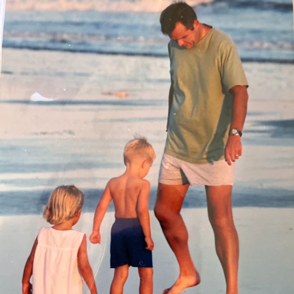 A man in a green shirt and shorts walks barefoot on the beach with two young children, a girl in a white dress and a boy in blue shorts, near the water’s edge.