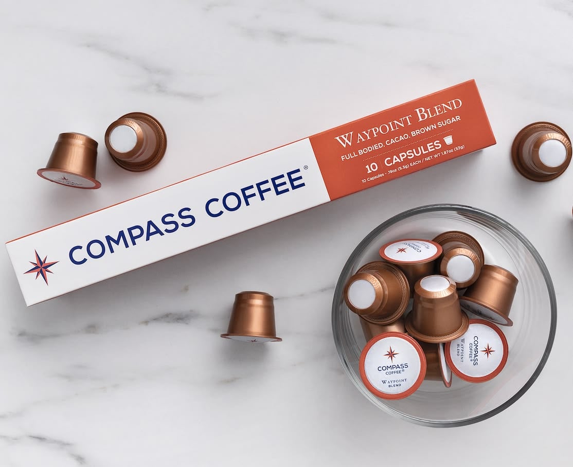 A box labeled "Compass Coffee Waypoint Blend" with several brown coffee capsules, some in a glass bowl and some scattered on a white marble surface. The box states it contains 10 capsules.