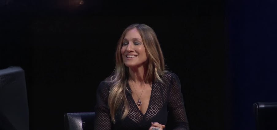 A woman with long, blonde hair, wearing a black patterned top and a necklace, sits and smiles with her eyes closed against a dark background.