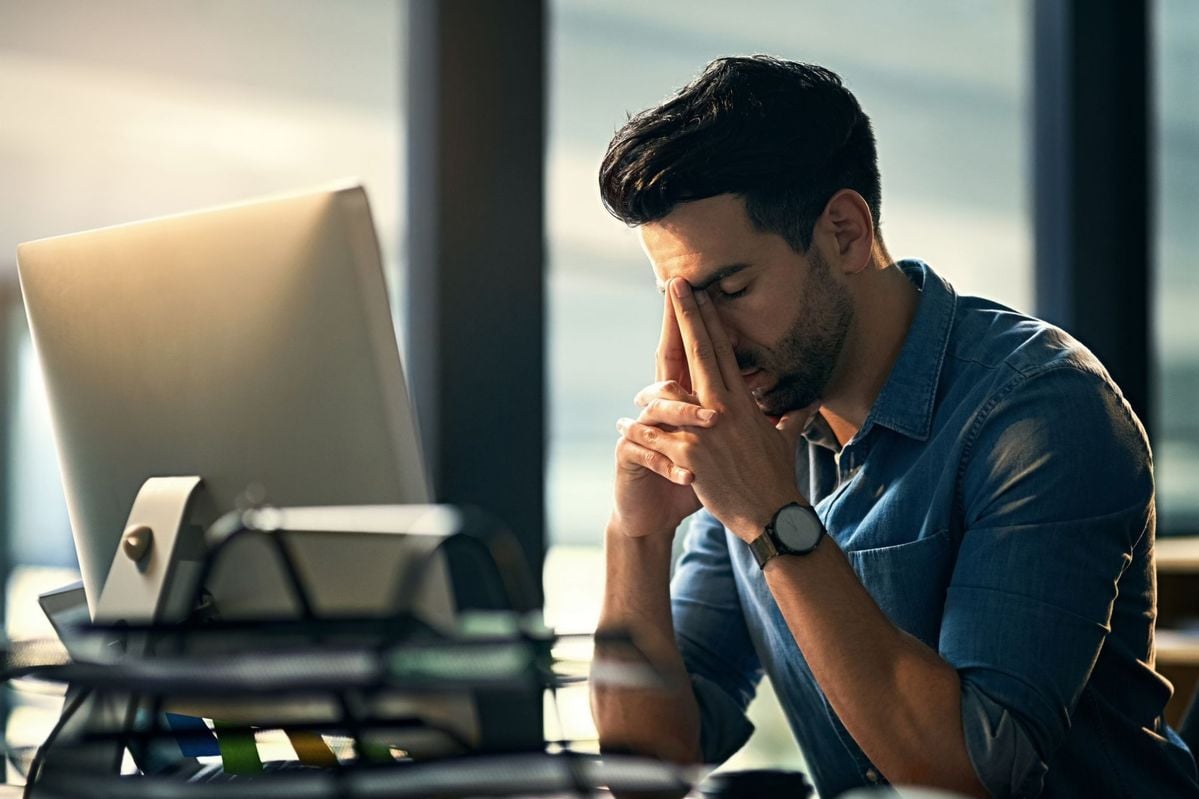 A man sits at a desk with his eyes closed and fingers pinching the bridge of his nose, appearing stressed or tired, in front of a computer in an office setting.