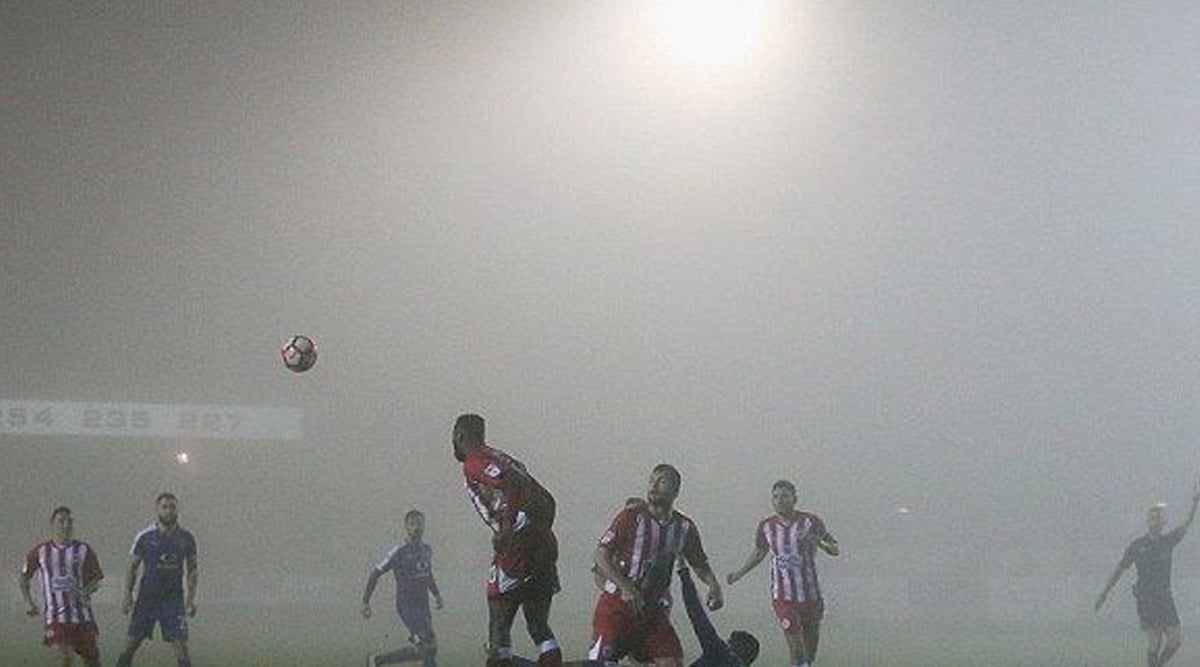 A group of soccer players in red and white and blue uniforms play a match in heavy fog, making the background and scoreboard barely visible under bright stadium lights.