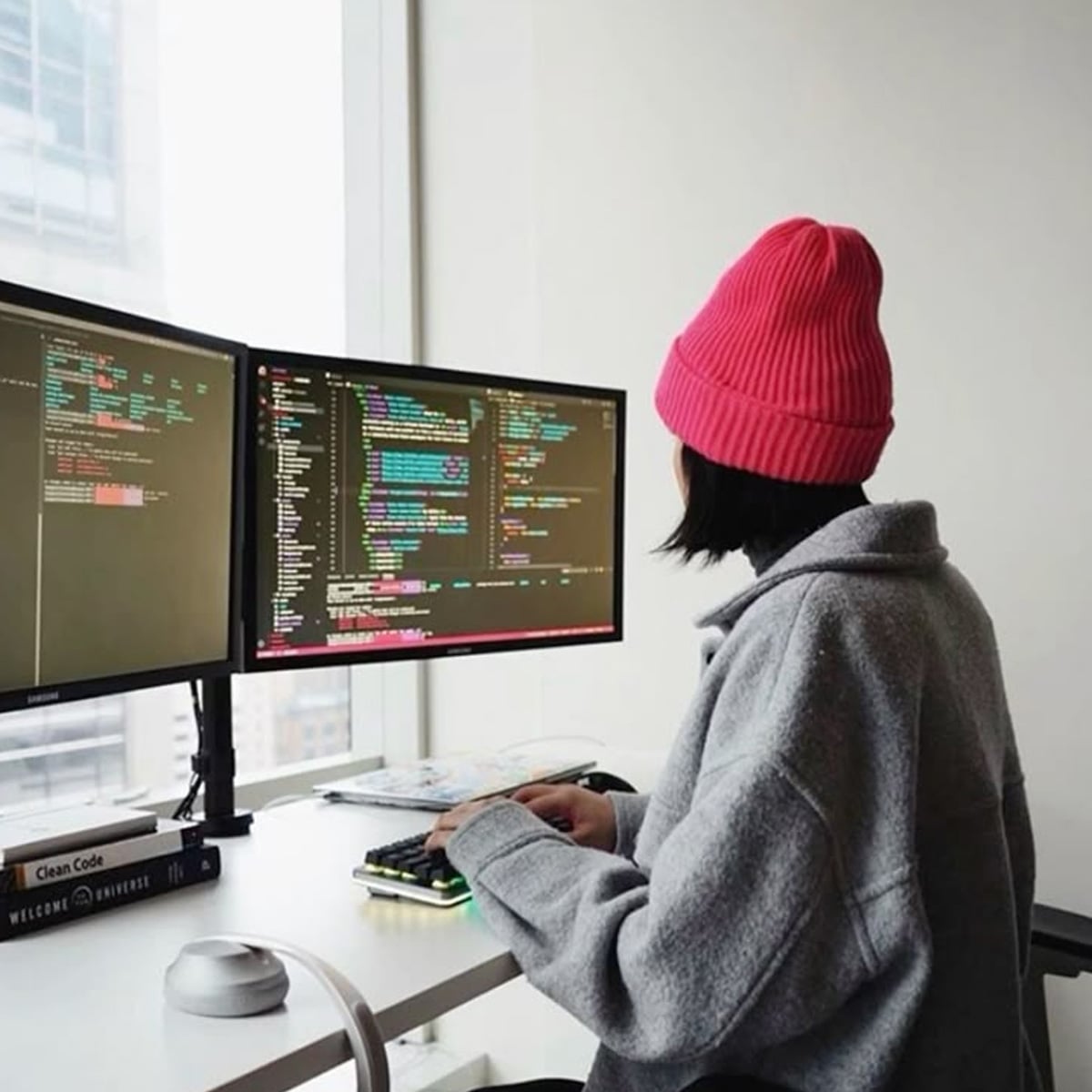 A person wearing a pink beanie and gray jacket works at a desk with two monitors displaying colorful code. The workspace is by a large window with city buildings visible outside.