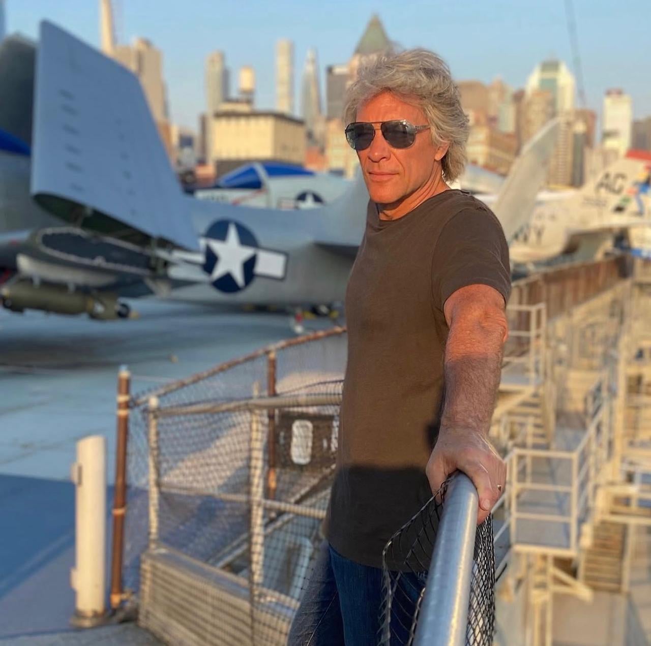 A man wearing sunglasses and a dark t-shirt leans on a railing aboard an outdoor aircraft carrier museum, with vintage military planes and a city skyline in the background.