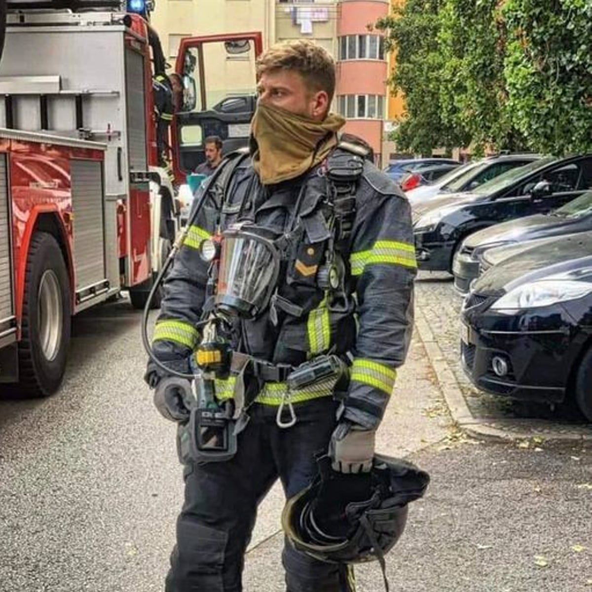A firefighter in full gear, holding a helmet and standing on a street near fire trucks and parked cars. Trees and buildings are visible in the background.