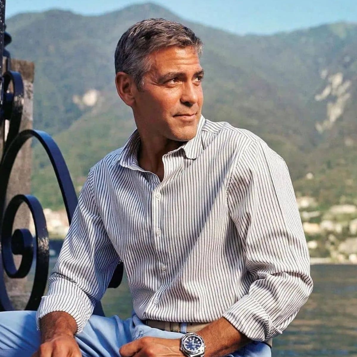 A man with gray hair wearing a light striped shirt and blue trousers sits outdoors by a lake, with green mountains and a village in the background on a sunny day.