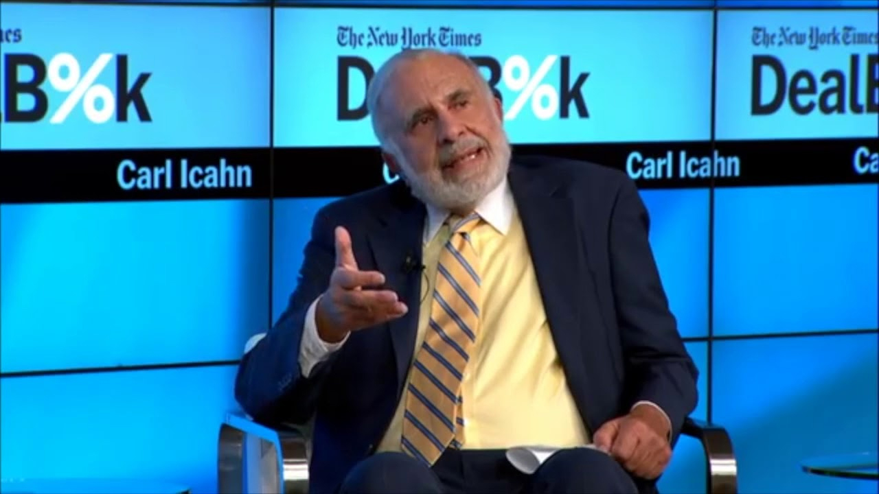 An older man in a suit and striped tie gestures while speaking at The New York Times DealBook event, with "Carl Icahn" displayed on the screen behind him.