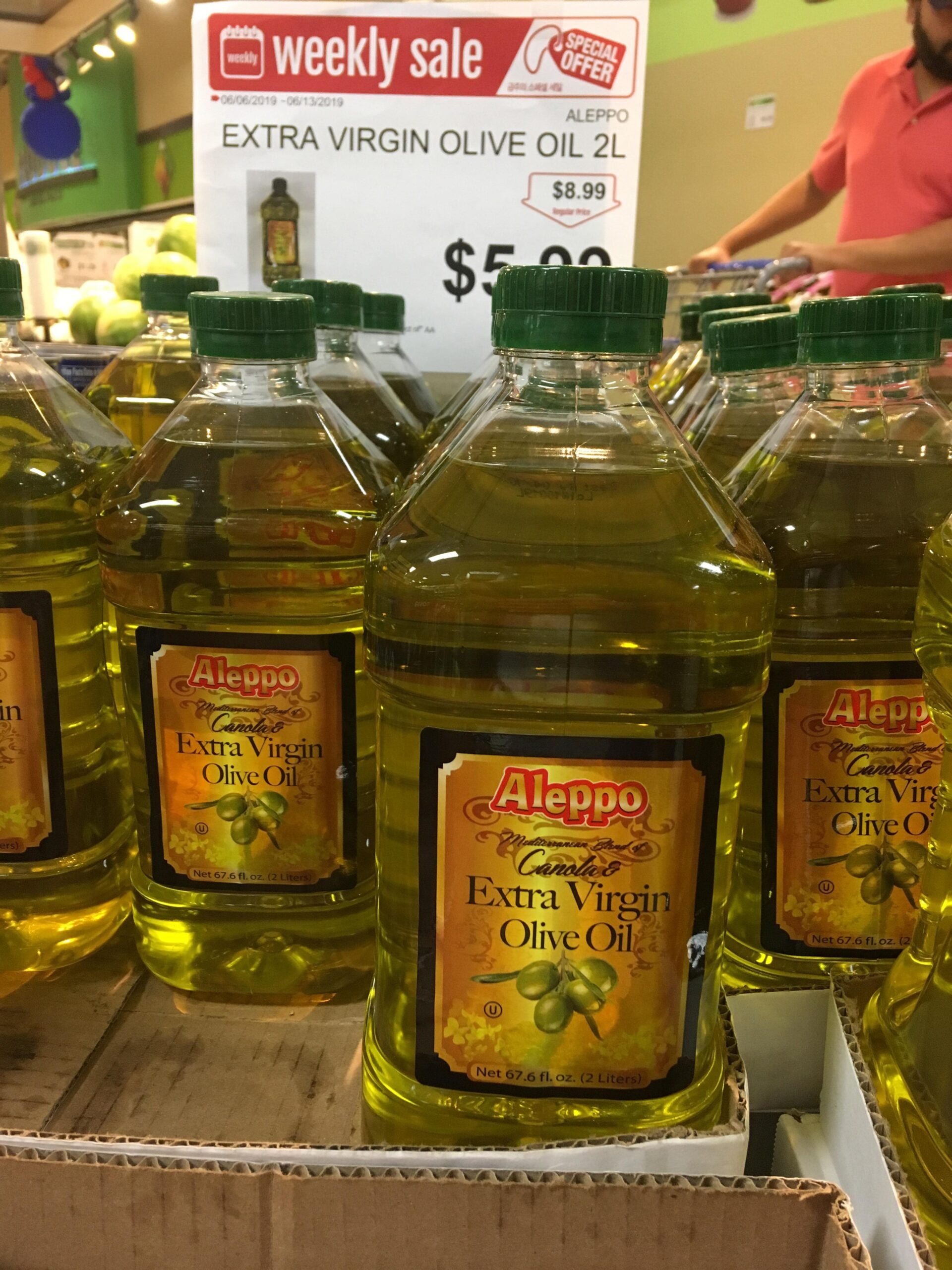 Bottles of Aleppo brand extra virgin olive oil are displayed on a store shelf under a weekly sale sign, priced at $5.99 each for 2 liters.