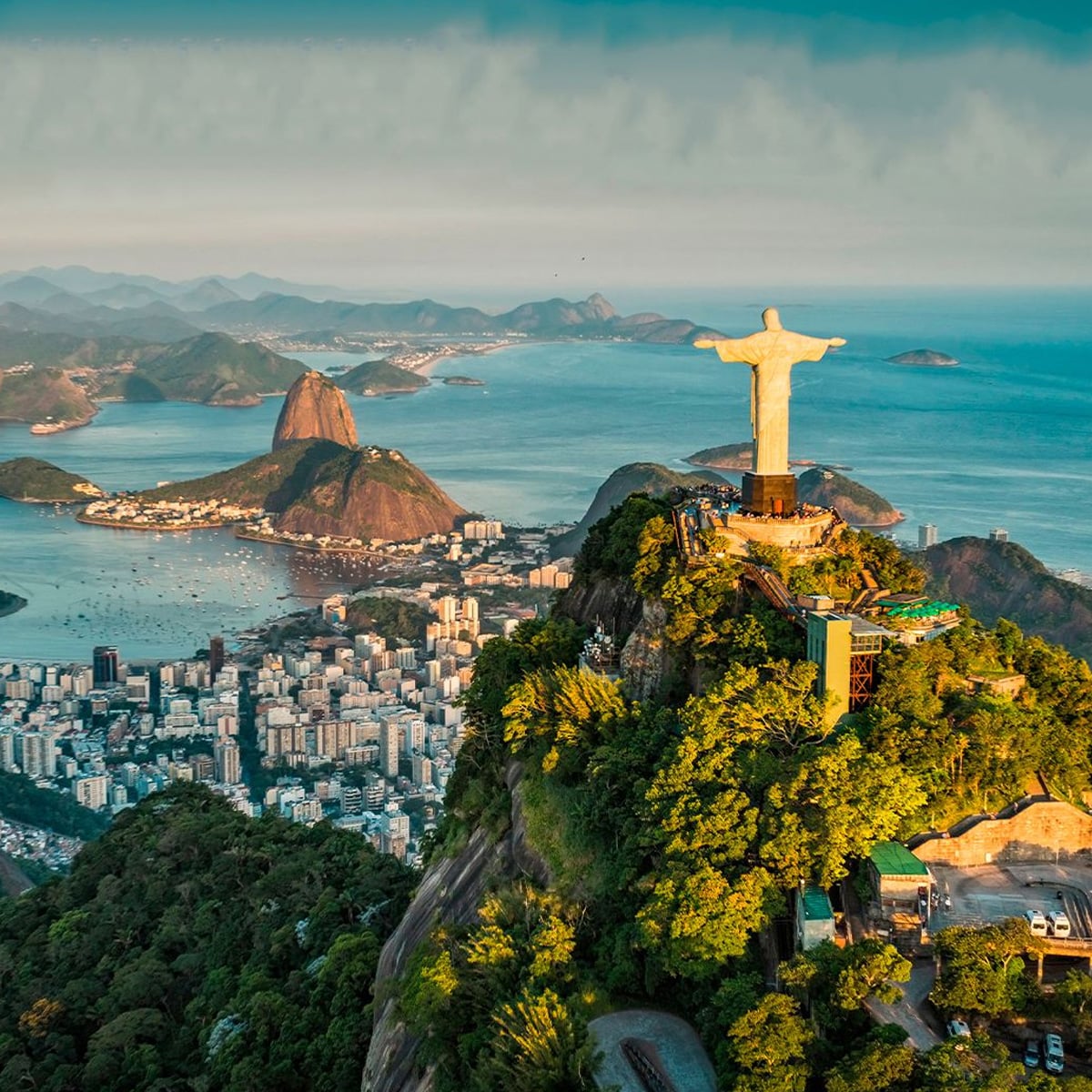 Aerial view of Rio de Janeiro showing the Christ the Redeemer statue atop a green mountain, with Sugarloaf Mountain, city buildings, and the ocean in the background under a blue sky.