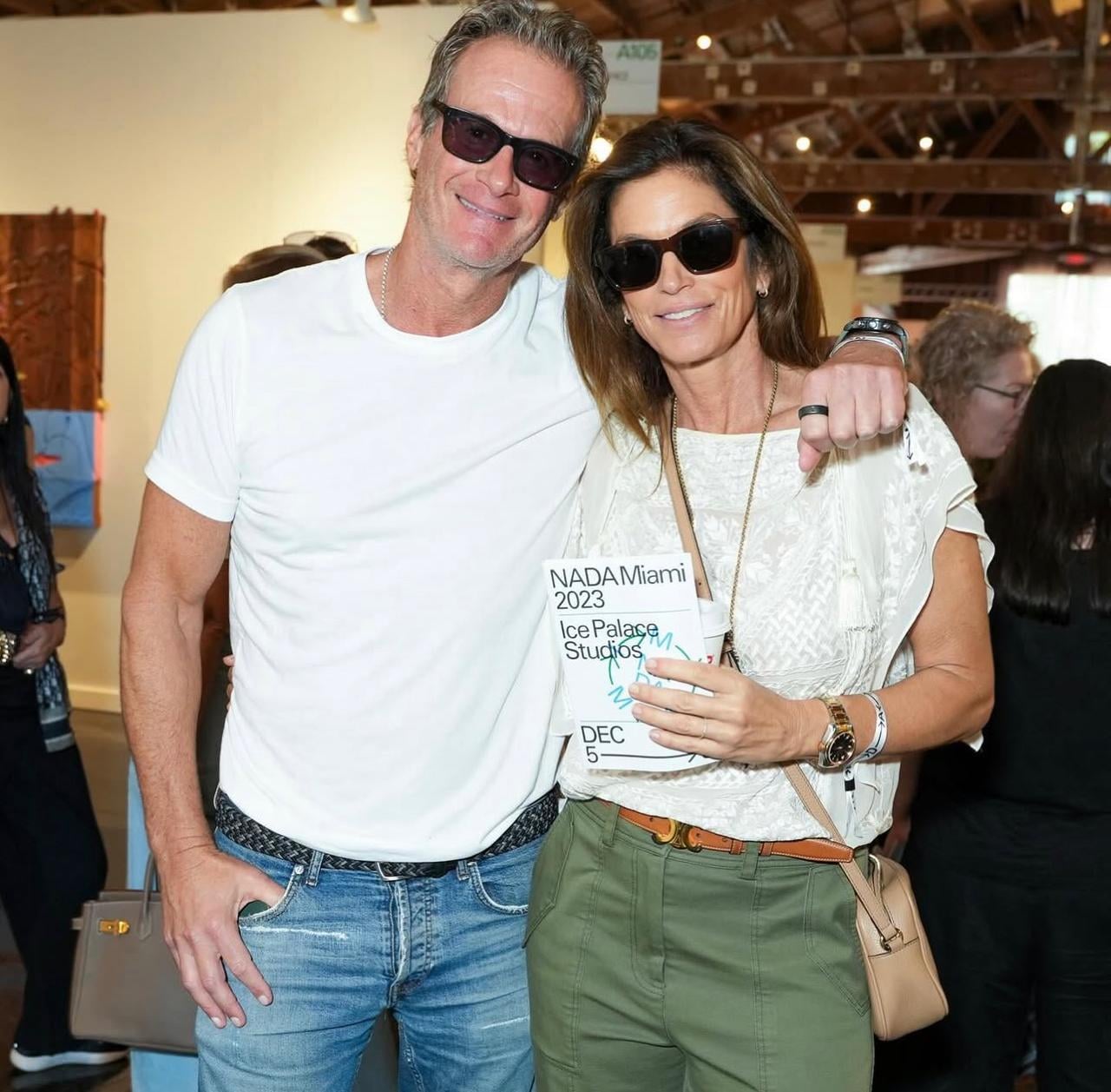 A smiling man and woman wearing sunglasses pose together at an indoor event. The woman holds a NADA Miami 2023 program. Both are casually dressed and stand close to each other in a well-lit gallery setting.